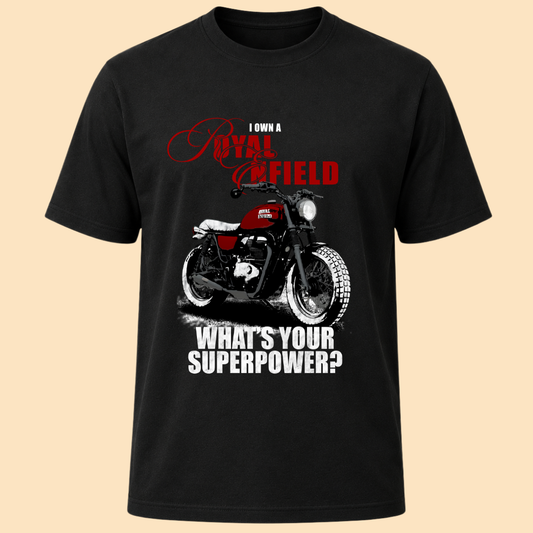 A black short-sleeve t-shirt with a graphic print of a red and black motorcycle, accompanied by the text 'I OWN A ROYAL ENFIELD' in red and 'WHAT'S YOUR SUPERPOWER?' in white, displayed against a solid beige background.