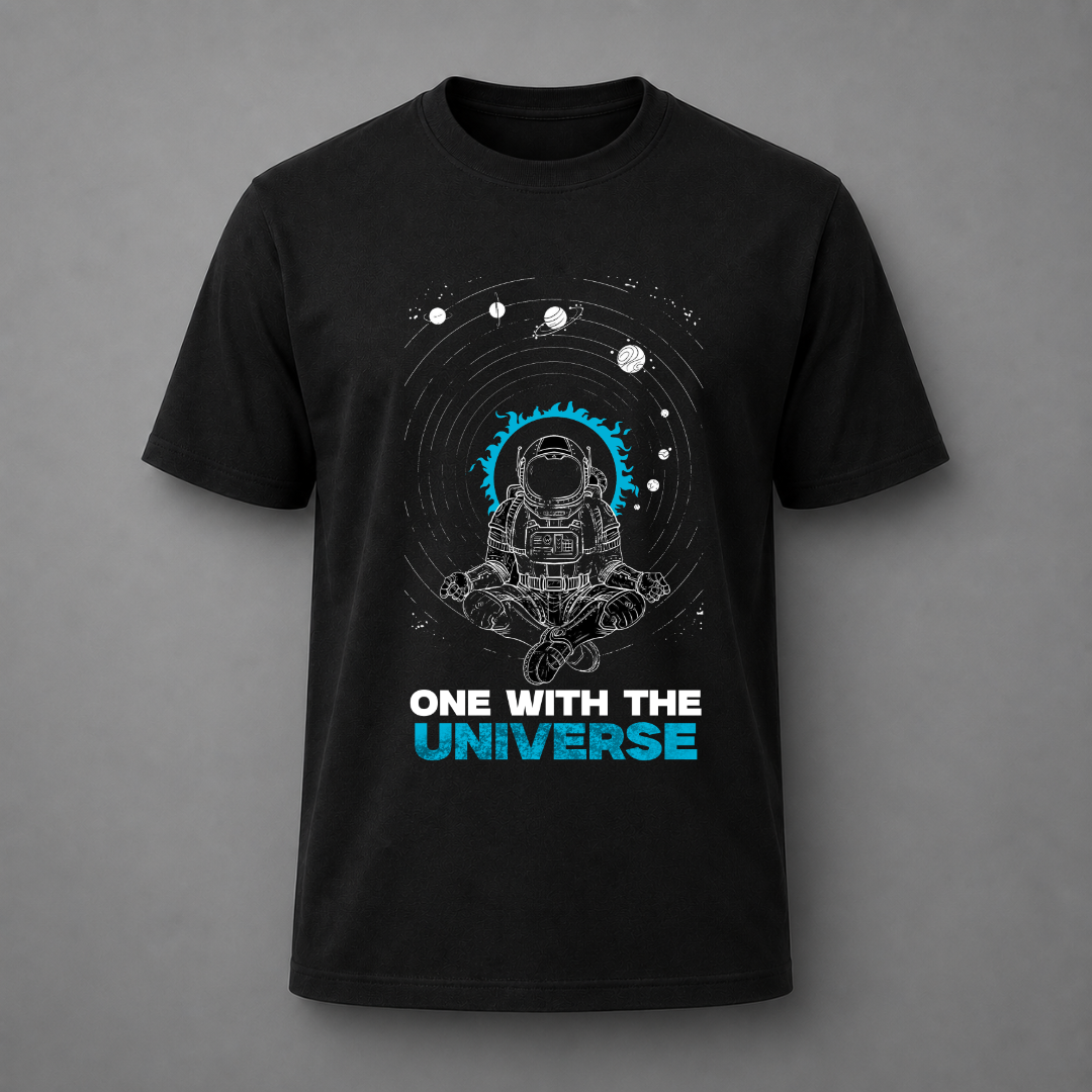 A black short-sleeve t-shirt with a graphic of an astronaut meditating inside a circular space design, with the text 'ONE WITH THE UNIVERSE' printed below in white and blue, displayed on a neutral gray background.
