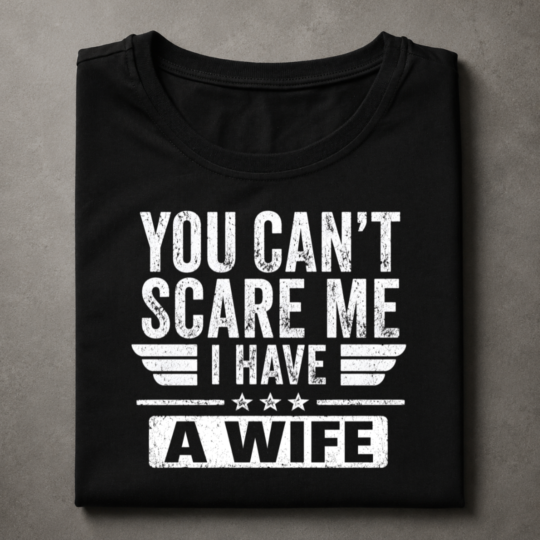 A folded black t-shirt with a distressed white graphic print that reads 'YOU CAN'T SCARE ME I HAVE A WIFE' with wing and star details, displayed on a textured gray background.