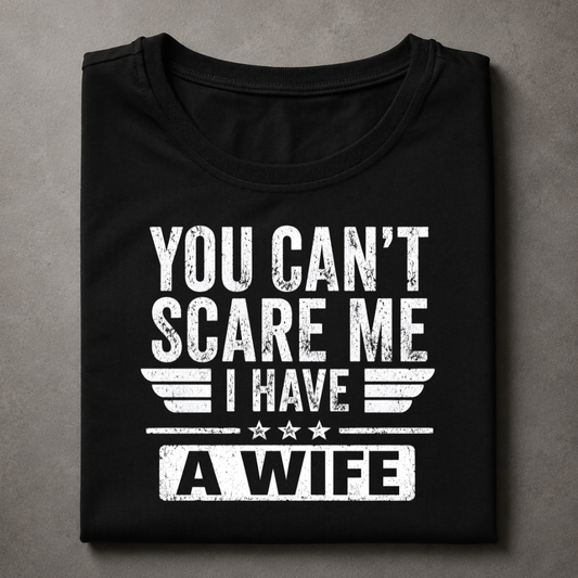 A folded black t-shirt with a distressed white graphic print that reads 'YOU CAN'T SCARE ME I HAVE A WIFE' with wing and star details, displayed on a textured gray background.