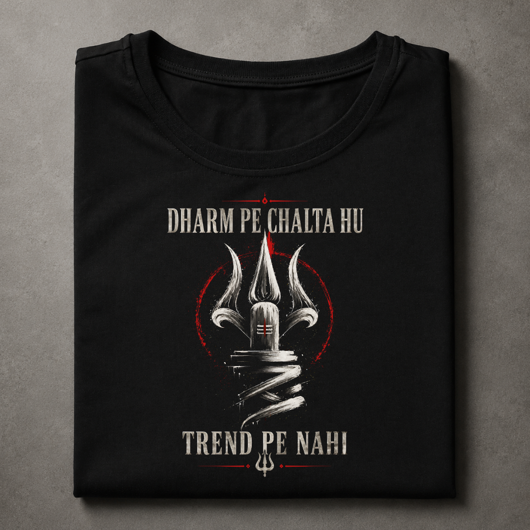 A folded black t-shirt with a graphic print featuring a trident, red circular accents, and white text reading 'DHARM PE CHALTA HU' and 'TREND PE NAHI', displayed on a textured gray background.