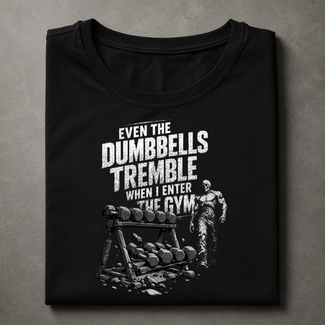 A folded black t-shirt with a distressed white graphic print featuring the text 'EVEN THE DUMBBELLS TREMBLE WHEN I ENTER THE GYM' and an illustration of a person next to a rack of dumbbells, displayed on a textured gray background.