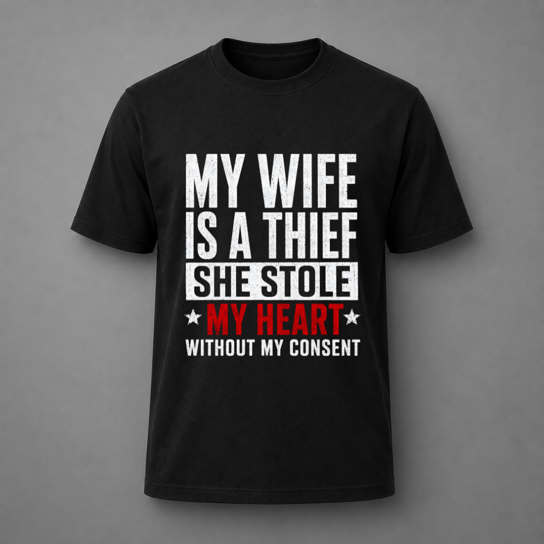 A black short-sleeve crewneck t-shirt with a humorous text graphic on the front. The graphic reads 'MY WIFE IS A THIEF SHE STOLE MY HEART WITHOUT MY CONSENT' in bold white and red lettering, displayed against a neutral gray background.