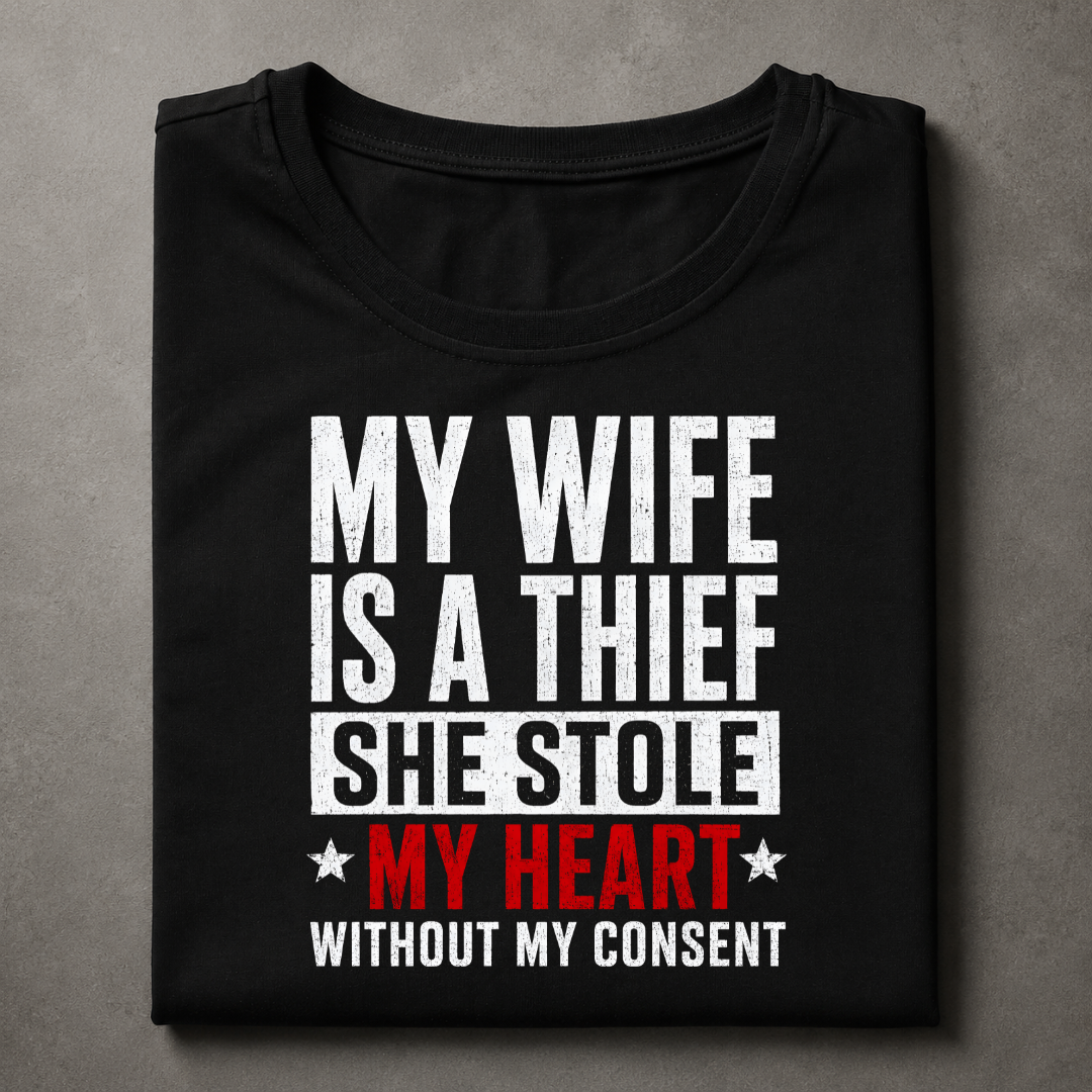 A folded black t-shirt with a humorous text graphic reading 'MY WIFE IS A THIEF SHE STOLE MY HEART WITHOUT MY CONSENT' in distressed white and red lettering, displayed on a textured gray background.