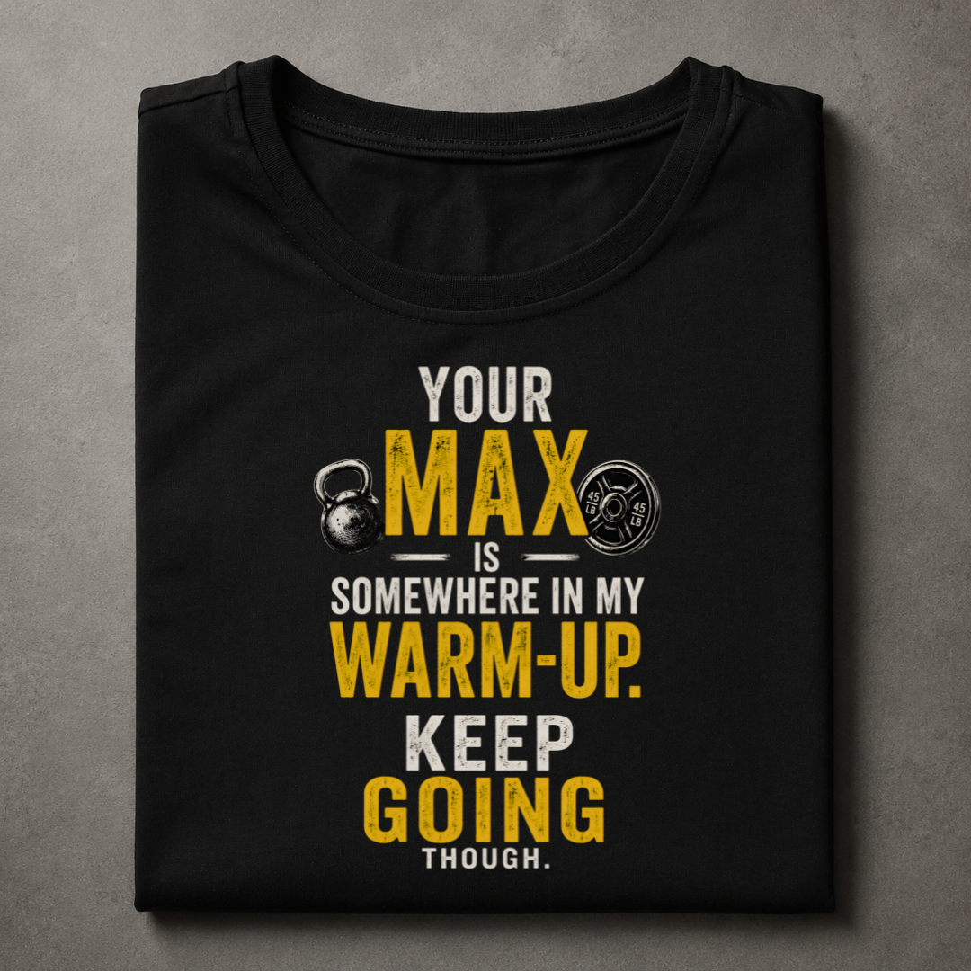 A folded black t-shirt with a humorous gym graphic printed on the front, displayed flat against a textured gray background. The graphic features the text 'YOUR MAX IS SOMEWHERE IN MY WARM-UP.